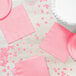 A table with a white cake and pink paper plates and napkins, with a pink napkin on the table with confetti.