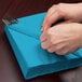 A person wrapping a turquoise blue Creative Converting luncheon napkin around a fork and knife.