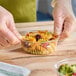 An 8 oz. clear round plastic deli container filled with pasta salad, being held by gloved hands on a wooden surface.