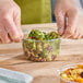 A clear 12 oz. round plastic deli container filled with broccoli salad, being held by gloved hands.