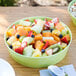 A bowl of fruit salad with a strawberry on top in a lime melamine bowl with a spoon on a napkin.