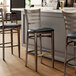 Three Lancaster Table & Seating Spartan Series Metal Ladder Back Bar Stools with Black Vinyl Seats on a counter.