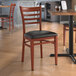 A Lancaster Table & Seating metal ladder back chair with mahogany wood grain finish and black vinyl seat at a table in a restaurant.