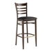 A Lancaster Table & Seating metal ladder back bar stool with dark walnut wood grain finish and black vinyl seat.