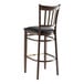 A Lancaster Table & Seating Spartan Series metal slat back bar stool with dark walnut wood grain finish and black vinyl seat.