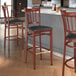 A group of Lancaster Table & Seating Spartan Series metal slat back bar stools with mahogany wood grain finish and black vinyl seats next to a counter.