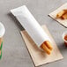 A churro in a white paper wrapper on a table with a cup of sauce and napkins.