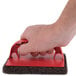 A close-up of a hand holding a red 3M Scotch-Brite Griddle Scrubber.