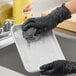 A person wearing black gloves is using a stainless steel scrubber to clean a soapy metal tray.