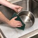 A person washing a metal pan with a 3M Scotch-Brite green scouring pad.