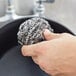 A hand holding a 3M stainless steel scrubber over a pan.