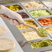 A person holding a Carlisle beige and red long handle spoon over food on a salad bar counter.
