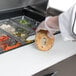 A hand in a glove holding a bagel over a counter with food in plastic containers.