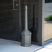 A gray Commercial Zone Smokers' Outpost cigarette receptacle next to a glass door.