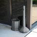 A grey Commercial Zone Smokers' Outpost cigarette receptacle with a black lid next to a glass door.