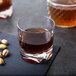 A GET SAN plastic rocks glass filled with brown liquid on a table next to nuts.