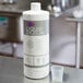 A white bottle of Urnex Dezcal liquid scale remover on a table next to a plastic measuring cup.