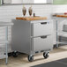 A stainless steel undercounter freezer with two drawers and caster wheels, shown in a commercial kitchen setting.