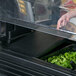 A hand opening a black Cambro well cover on a counter with containers of greens.