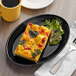 A Tuxton black oval china platter with food on a wood table and a yellow mug with black liquid.