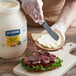 A gloved hand spreads Hellmann's Extra Heavy Mayonnaise on a sandwich.