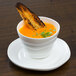 A white 10 Strawberry Street porcelain cup filled with orange soup with toast on top.