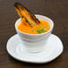 A white cup with a 10 Strawberry Street Ripples bright white saucer on a table with orange soup and a piece of toast on top.