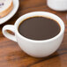 A Tuxton Nevada eggshell china cup filled with coffee on a wooden table.