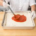A person using a spoon to spread red sauce on pizza dough in an American Metalcraft aluminum cake pan.