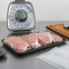 A black CKF foam meat tray of raw meat on a counter next to a cutting board with a piece of meat on it.