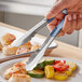 A person using Vollrath stainless steel tongs with blue Kool-Touch handles to serve food on a plate.