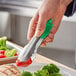 A person using Vollrath stainless steel tongs with a green handle to serve broccoli.