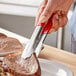 A pair of Vollrath stainless steel tongs with a red Kool-Touch handle holding a piece of meat.