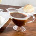 A Fineline clear plastic gravy boat on a table with brown liquid in it.