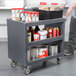 A man using a Cambro granite gray plastic service cart to store food containers and cans.