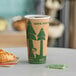 A close-up of a brown EcoChoice paper hot cup with green trees on it on a table with a cup of coffee and a croissant.