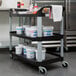 A black Rubbermaid utility cart with three shelves holding white buckets of food.