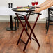 A person using an American Metalcraft mahogany wood tray stand to hold a tray with food and wine.