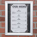 An 11" x 14" satin aluminum Aarco snap frame holding a menu on a brick wall.