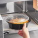 A hand using a Choice black plastic container to heat food in a microwave.