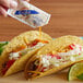 A hand holding a Crystal hot sauce packet pouring sauce onto two tacos on a table.