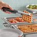 A hand using Vollrath Kool-Touch Pom Tongs to grab a waffle from a tray of food.