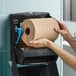 A large roll of natural kraft hardwound paper towel being loaded into a black wall-mounted dispenser.