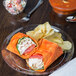 A Choice Crystal clear plastic plate with a sandwich wrapped in orange paper and a bowl of food on a wood table.