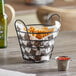 A round black wire basket with handles holding a basket of fries with a bottle of ketchup on a table.