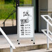 An Aarco white aluminum A-frame sign board with black writing on it outside of a building.