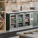 A stainless steel underbar height back bar refrigerator with three glass doors, stocked with various beverages.