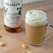 A glass mug of Monin Macadamia Nut syrup-flavored coffee with whipped cream and nuts next to a bottle of Monin Macadamia Nut syrup.