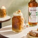 A glass mug of iced coffee topped with whipped cream and caramel, with a bottle of Monin Premium Macadamia Nut Flavoring Syrup in the background.