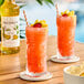 A bottle of Monin Premium Mango Flavoring Syrup next to two tall glasses of a bright orange tropical drink garnished with fruit and a cherry.